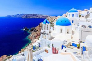 An island with white mosque shaped houses with blue rounded tops next to a blue sea