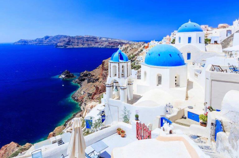 An island with white mosque shaped houses with blue rounded tops next to a blue sea